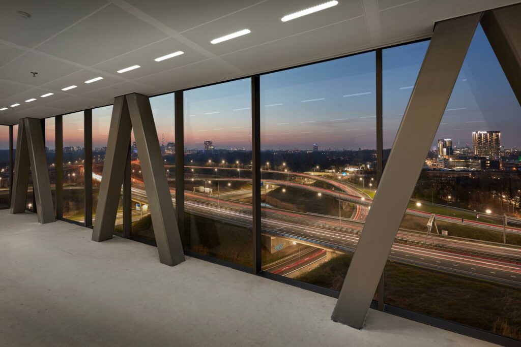 Uitzicht vanuit een modern kantoorgebouw aan de Joan Muyskenweg op een knooppunt van snelwegen en de skyline van Amsterdam bij schemering.